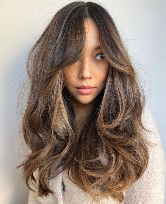 a long dark brunette hairstyle with caramel and golden blonde balayage, with bottleneck bangs and a lot of dimension and waves