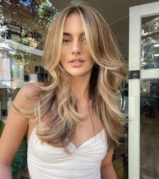 a lovely butterfly haircut on bronde hair, with blonde highlights and money piece, with feathered layers and a bit of waves is very cic